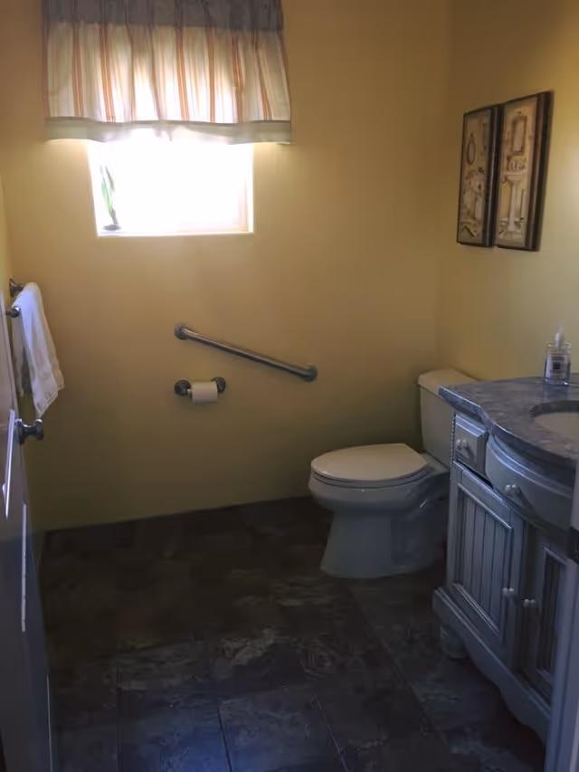 A small bathroom with a toilet, a vanity with a sink, and a window with a curtain. There is a grab bar on the wall next to the toilet, a toilet paper holder, and two framed pictures hanging on the wall above the toilet. The floor is tiled with dark tiles.