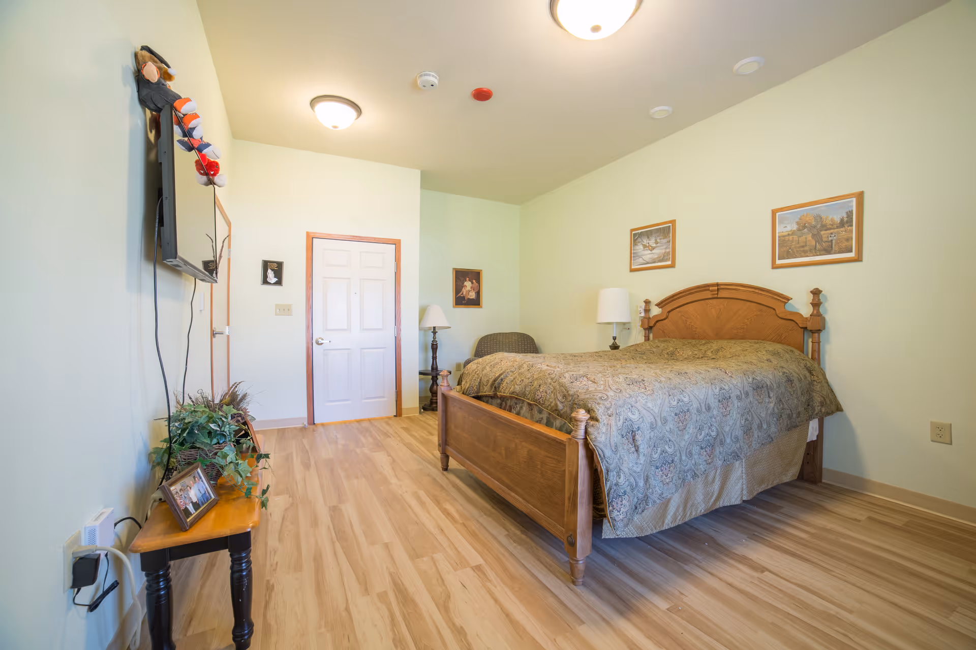 A furnished bedroom with a wooden bed, nightstand, wall-mounted TV and light green walls in an assisted living facility.