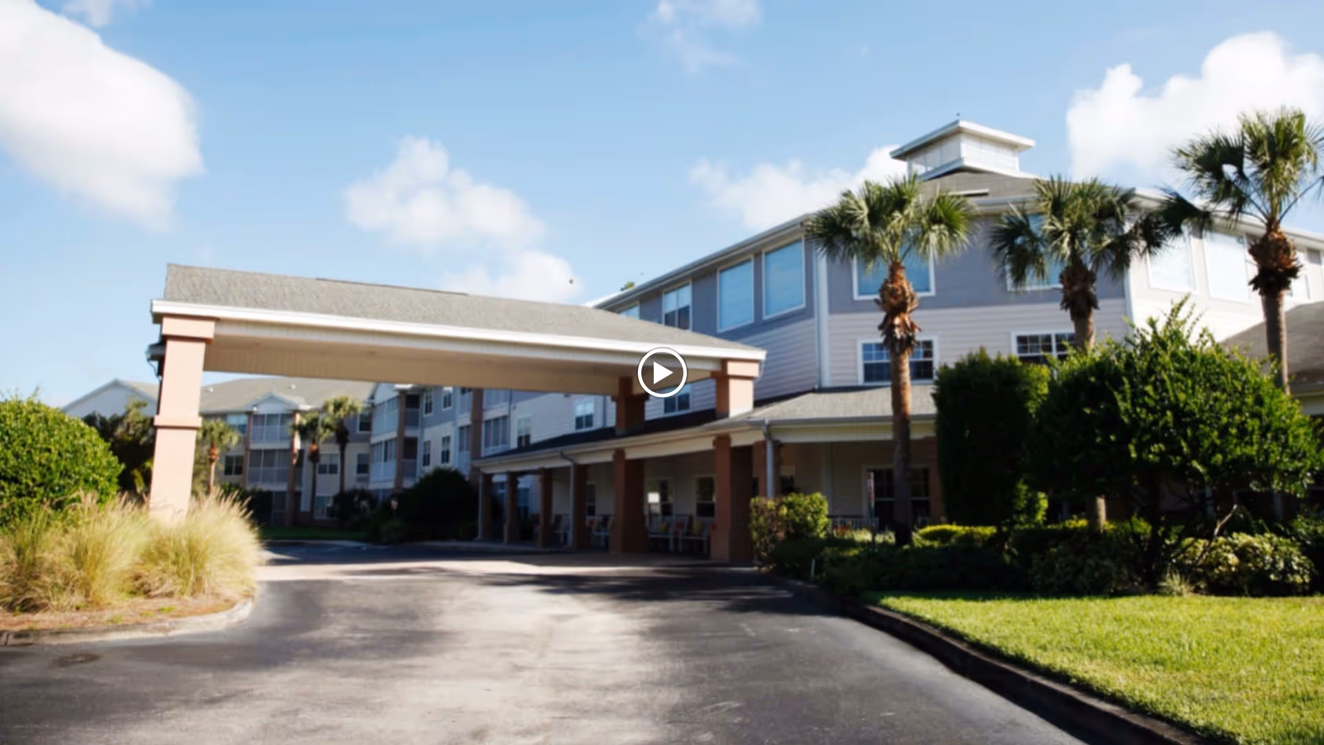 Exterior view of Addington Place of East Lake senior living facility showing a multi-story building with a covered entrance, palm trees, bushes, and a clear blue sky.