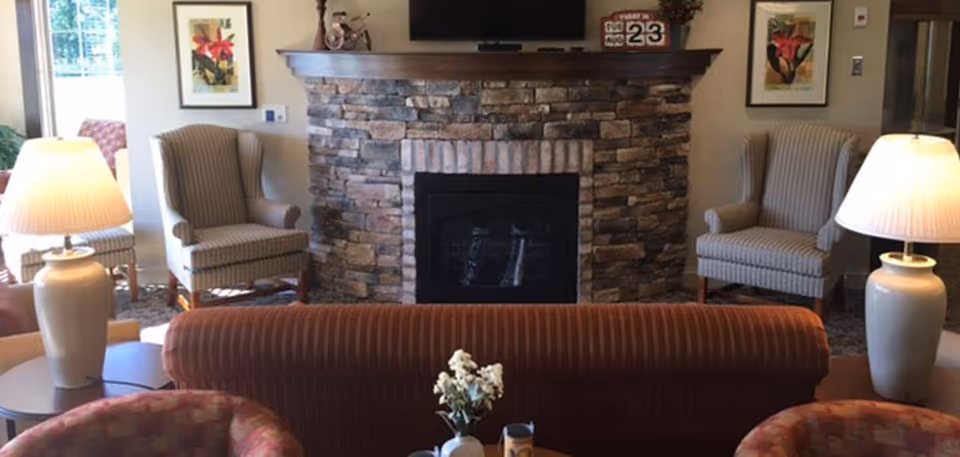 Seating area with a stone fireplace centered between two striped armchairs, sofas, a small table with flowers, and table lamps.