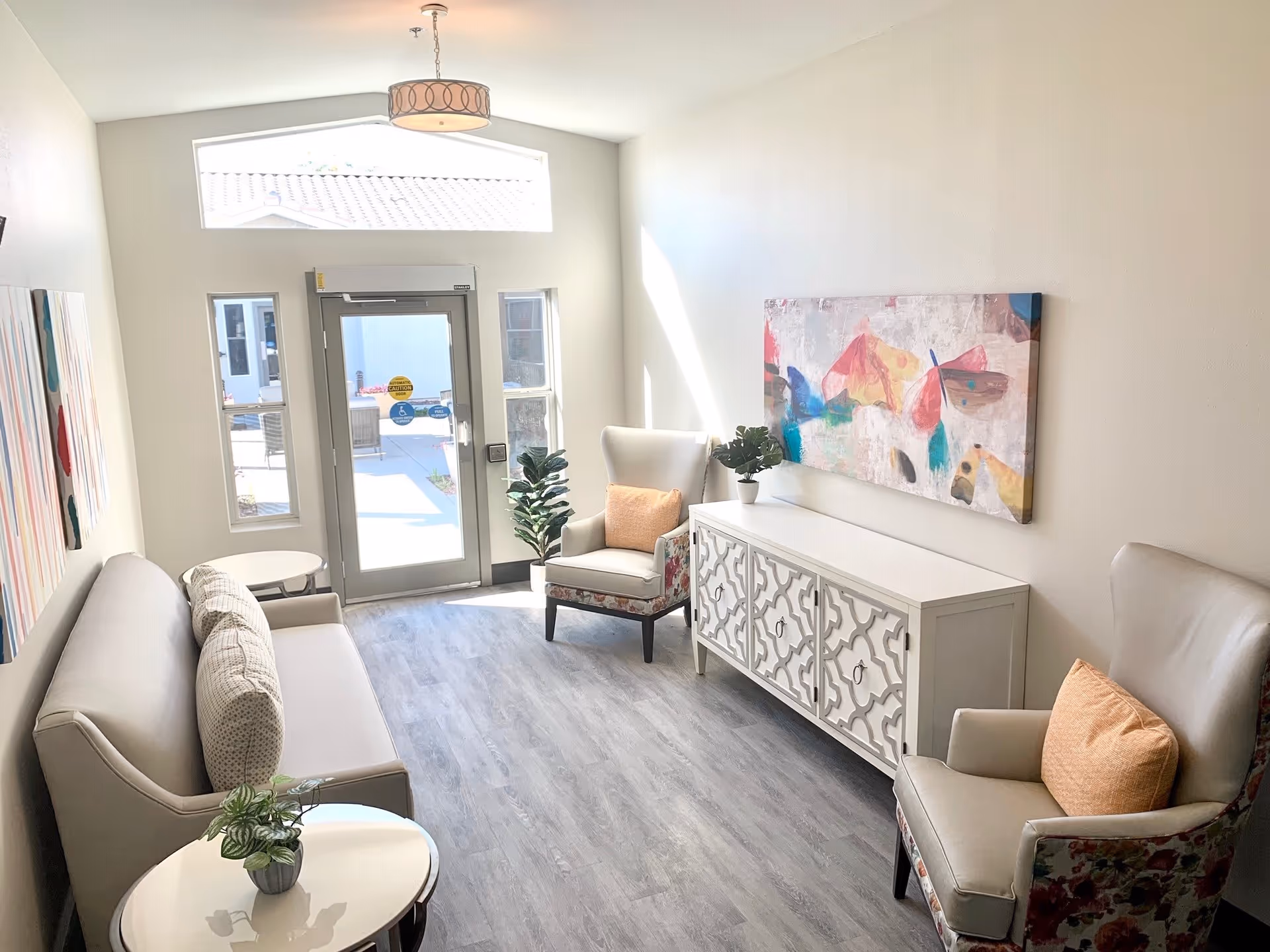 A bright and modern sitting area with a gray sofa, two armchairs with orange cushions, a white decorative cabinet, and colorful abstract artwork on the wall. There is a glass door and windows letting in natural light, and a small round table with a plant on it.