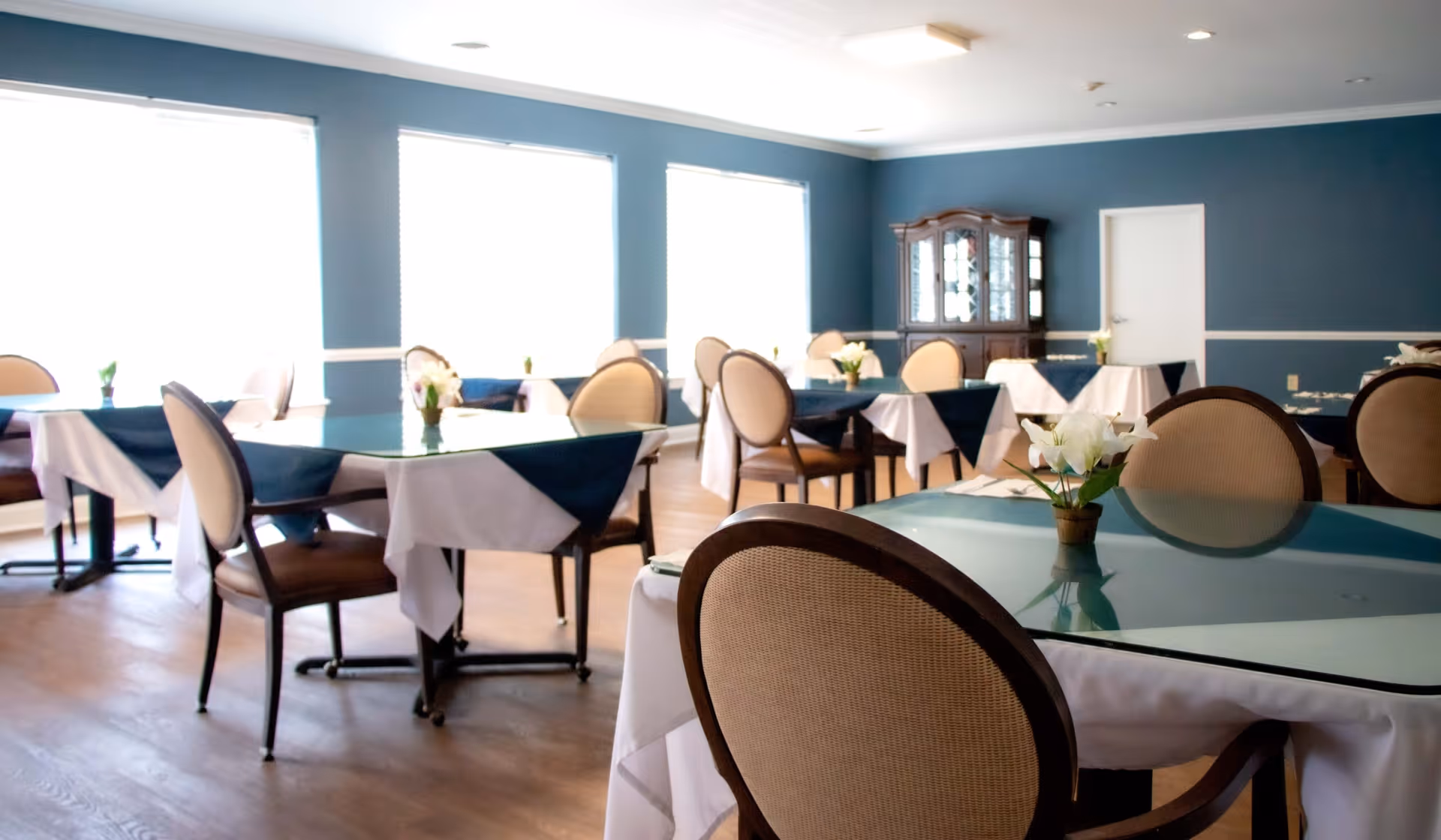 A dining room with multiple tables covered with white and blue tablecloths, each table decorated with a small flower arrangement. The room has blue walls, large windows letting in bright natural light, and a wooden china cabinet against the far wall.
