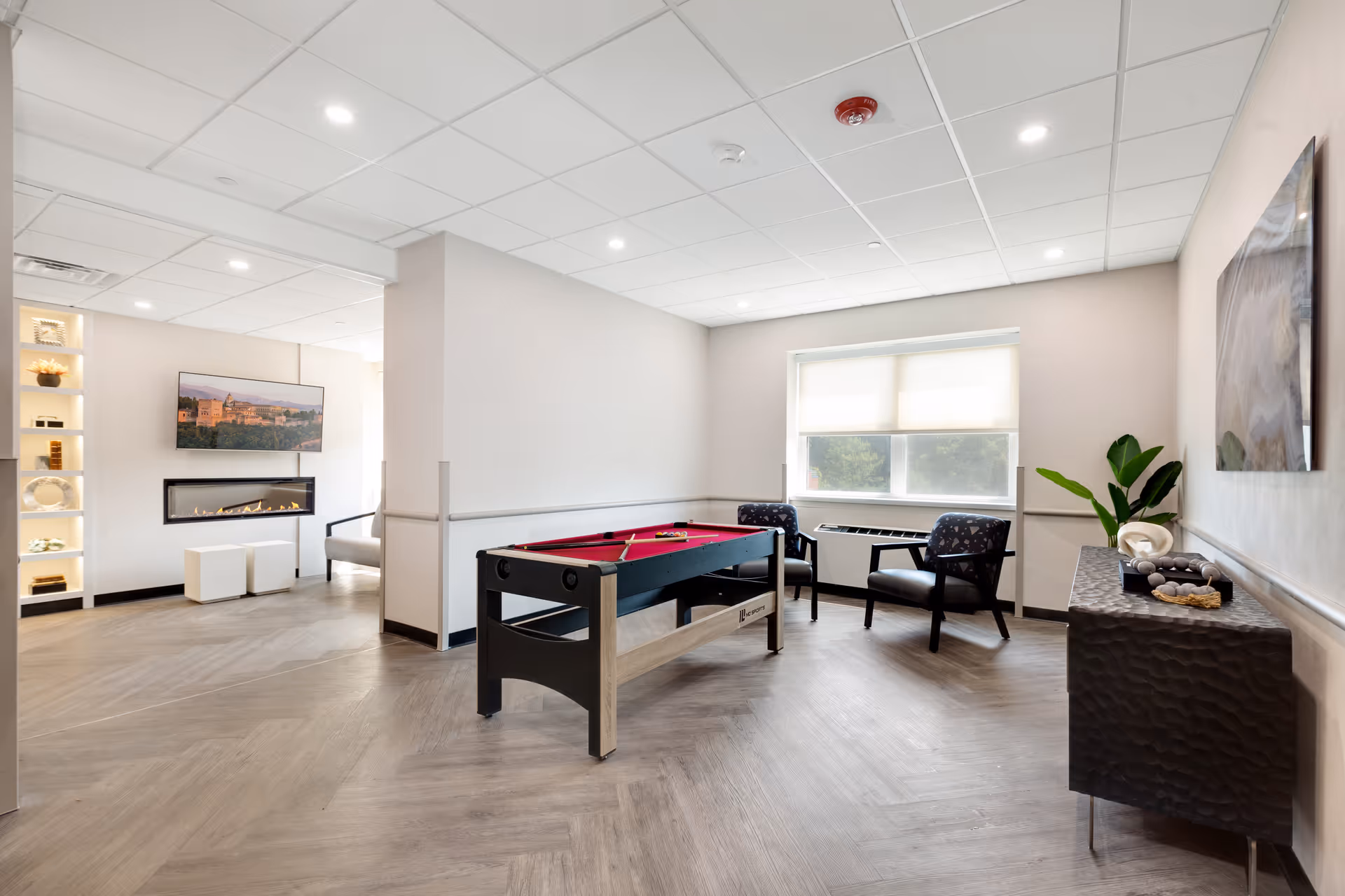 A bright and modern common area in a healthcare facility featuring a red felt pool table in the center, two upholstered chairs near a window with blinds, a decorative console table with a plant and ornaments, and a wall-mounted TV above a sleek electric fireplace in the adjacent room.