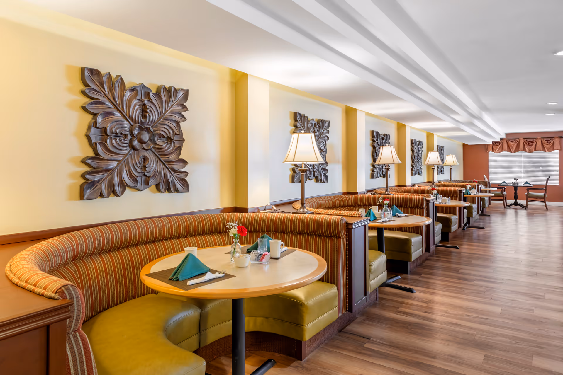 Bright dining room with curved striped booth seating, round tables set with napkins and lamps along a long wood‑floored room.
