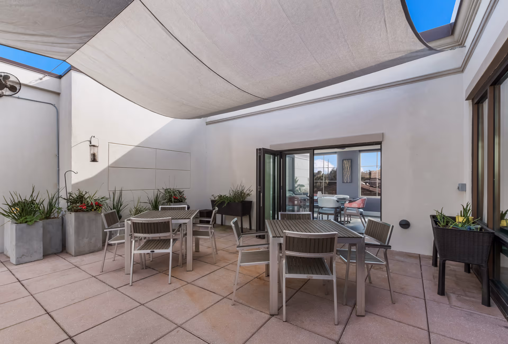 Outdoor patio area with several tables and chairs arranged on a tiled floor. The space is partially shaded by a large fabric canopy overhead. There are multiple planters with green plants and flowers along the walls. Glass doors open into an interior room with additional seating visible inside.