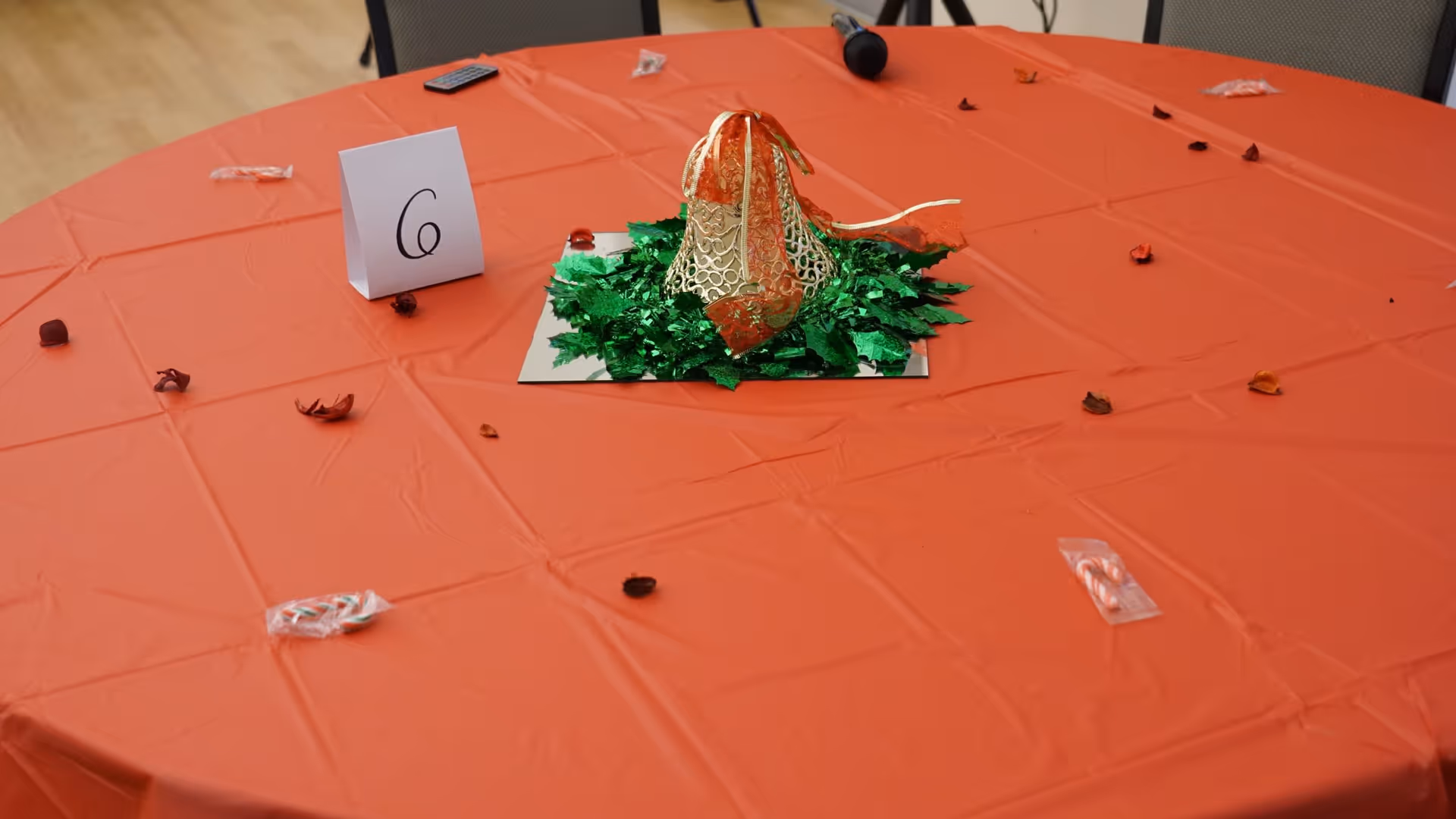 A round table covered with an orange tablecloth, decorated with a centerpiece featuring a gold and orange decorative bell surrounded by green foil leaves. The table also has scattered small wrapped candies and dried flower petals, a folded card with the number 6, and a microphone resting on the table. Chairs with mesh backs are visible around the table.