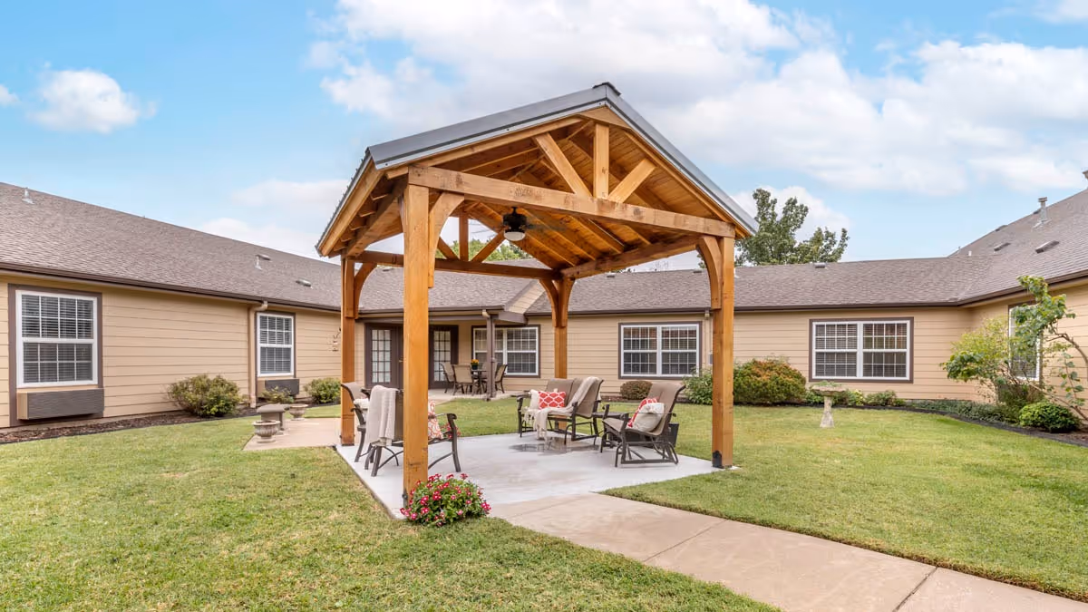 Outdoor courtyard area at Brookdale Bartlesville North featuring a wooden gazebo with seating underneath, surrounded by a well-maintained lawn and beige buildings with multiple windows.