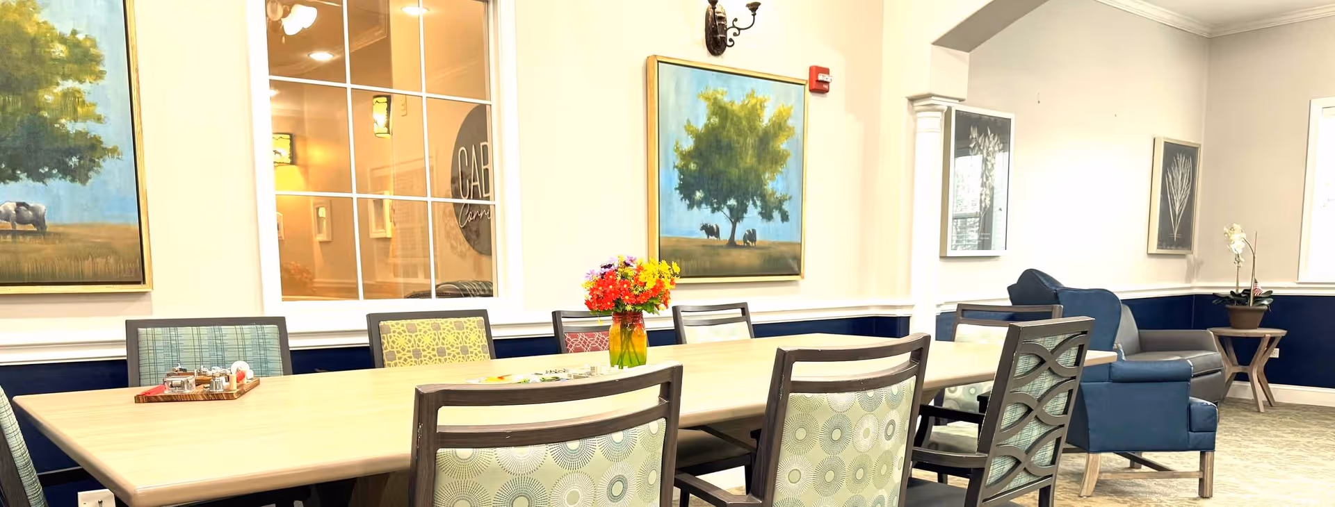 A bright and spacious dining area with a long wooden table surrounded by patterned chairs. A vase with colorful flowers is placed in the center of the table. The walls are decorated with framed paintings of trees and animals. In the background, there is a blue armchair and a small side table with a potted plant near a window.