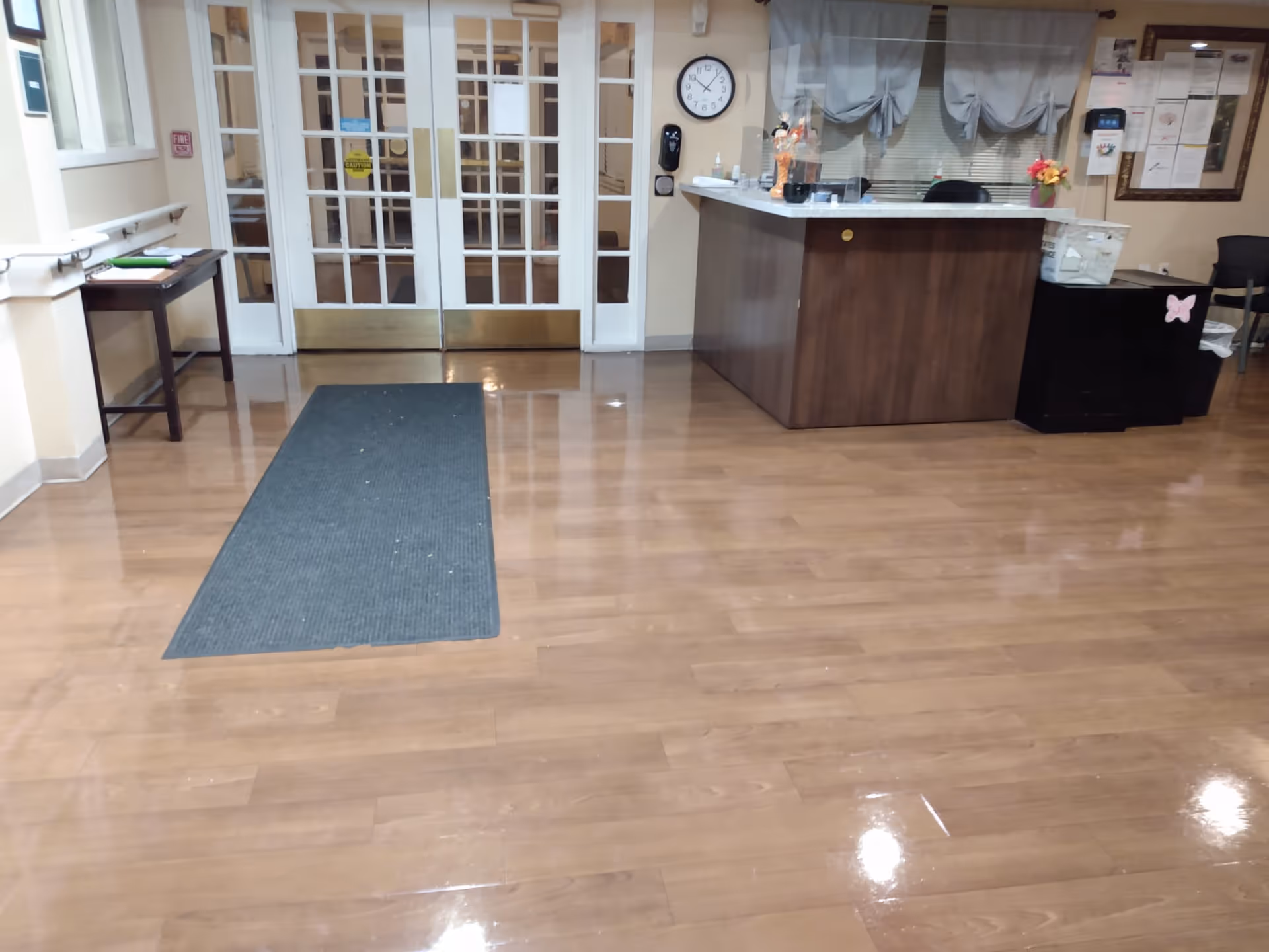 Reception area in a senior living facility with a wooden floor, a dark wooden reception desk on the right, a clock on the wall behind the desk, a small table with items on the left, and double glass doors in the background.