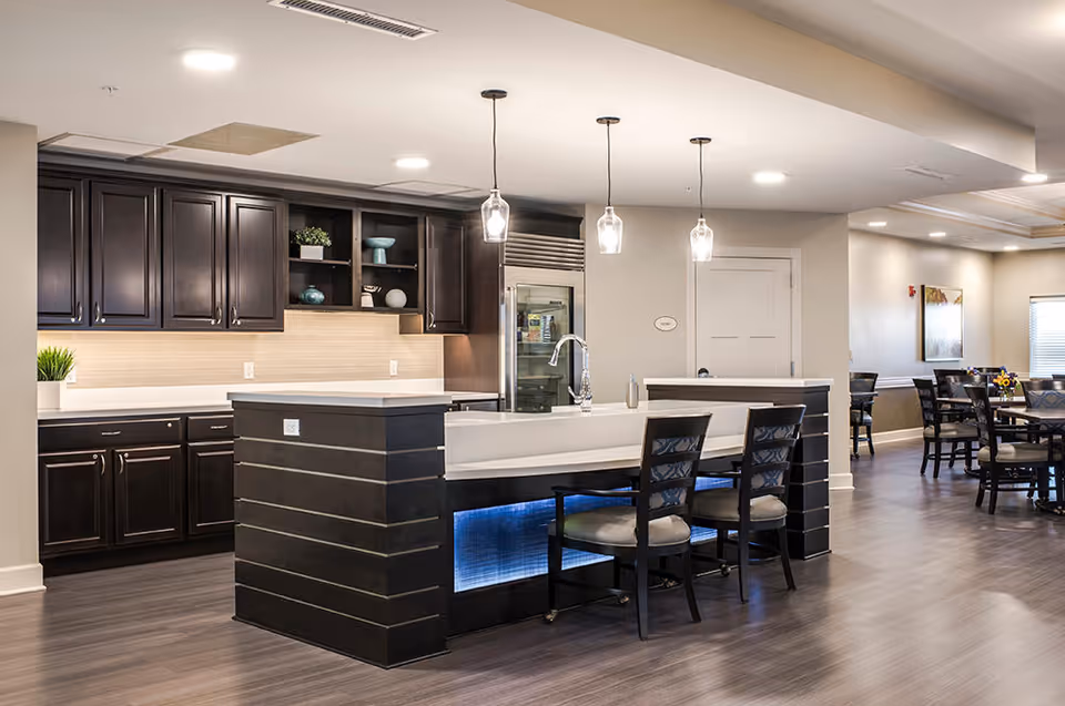 Modern kitchen area in a senior living facility with dark wood cabinets, a large island with a sink, three pendant lights hanging above, and two chairs at the island. In the background, there is a dining area with tables and chairs, light-colored walls, and wood flooring throughout.