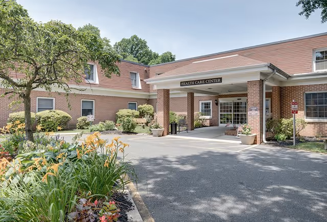 Exterior view of a brick health care center building with a covered entrance, surrounded by greenery and flowering plants under a clear sky.