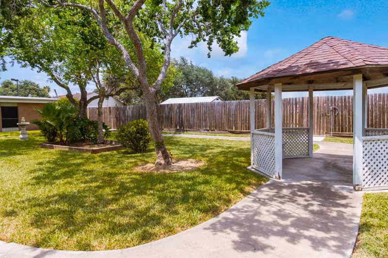 Outdoor garden area with green grass, trees, and a wooden gazebo with a shingled roof. A concrete pathway leads to the gazebo, and a wooden fence encloses the area under a clear blue sky.