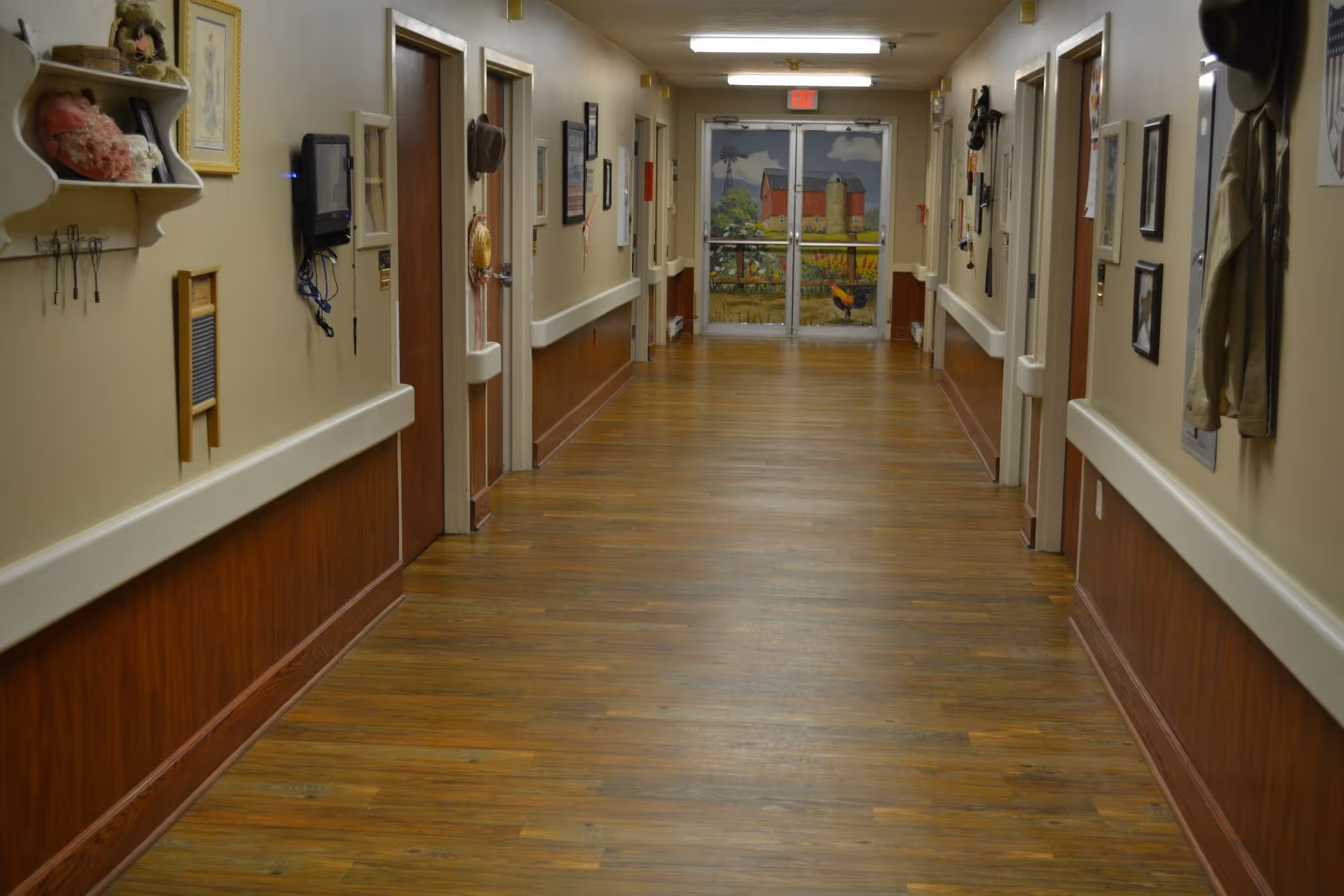Long interior hallway with wood floors, handrails and doors on both sides and a painted exit door at the far end.