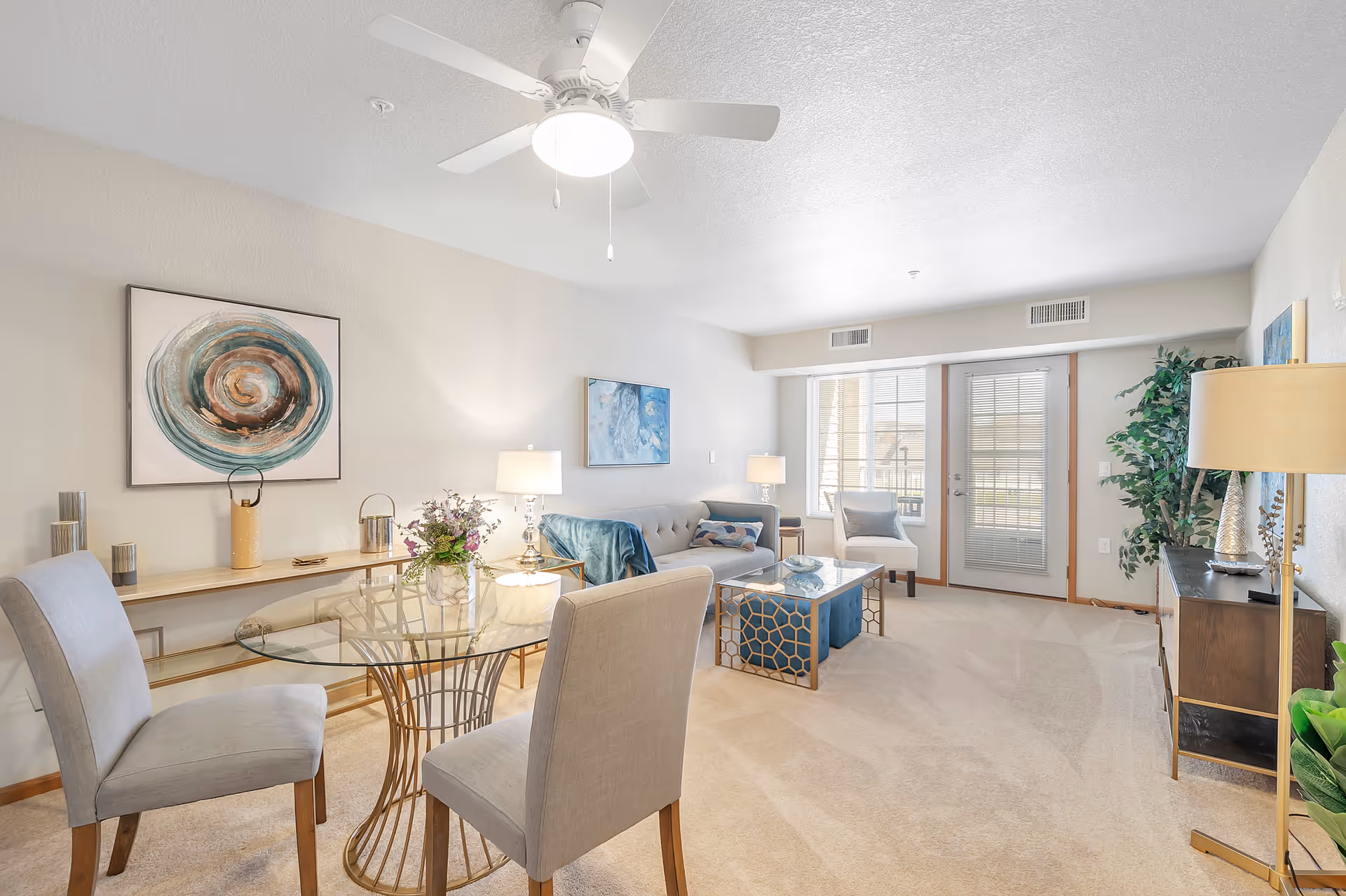 A bright and spacious living room with beige carpet and light-colored walls. The room features a glass-top dining table with two upholstered chairs, a beige sofa with blue and patterned cushions, a glass coffee table with a blue ottoman underneath, and a white armchair near a door with window blinds. The room is decorated with abstract wall art, table lamps, a ceiling fan with light, and several plants.