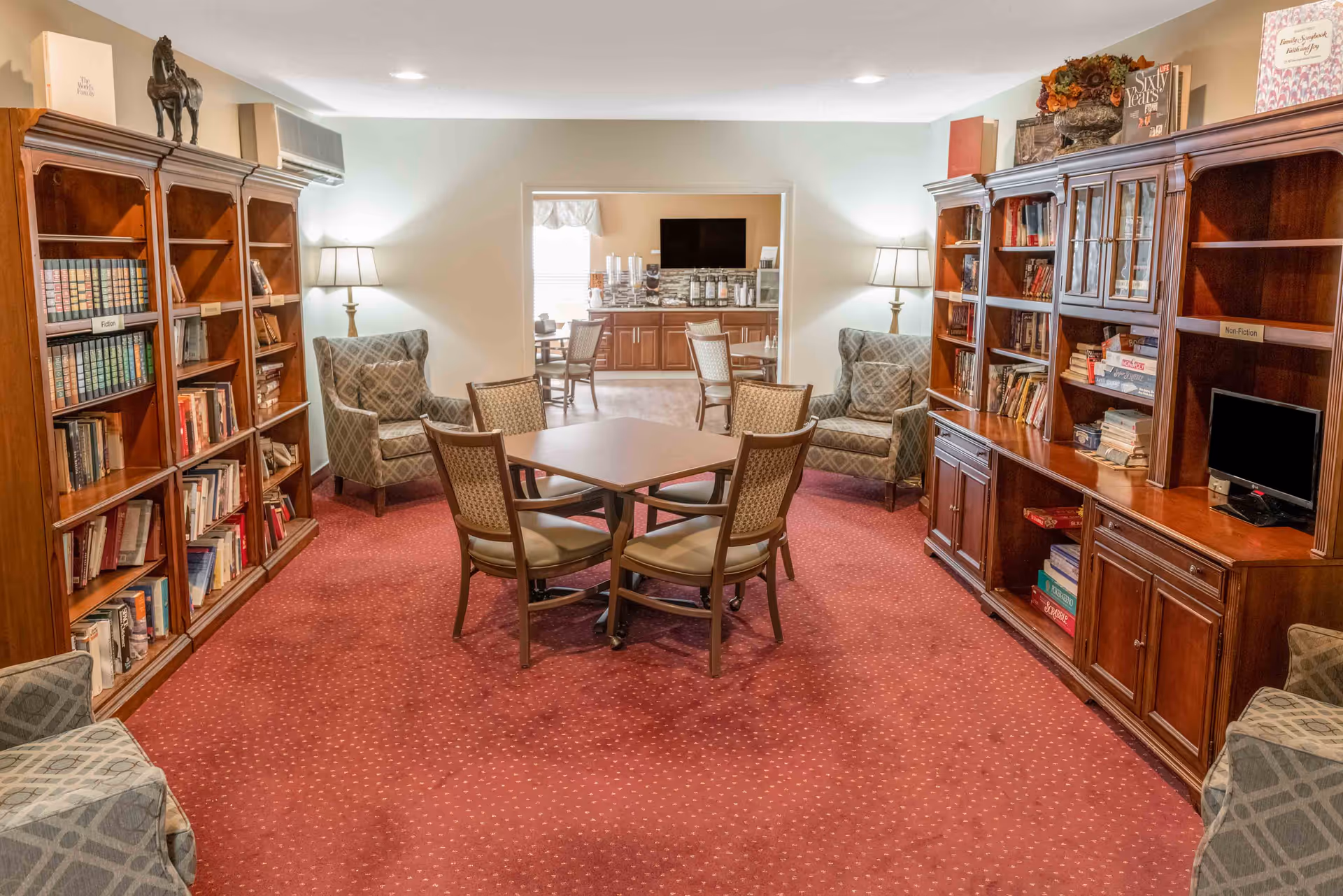 Cozy communal lounge/library with bookshelves, armchairs, and a central table in a senior living facility.