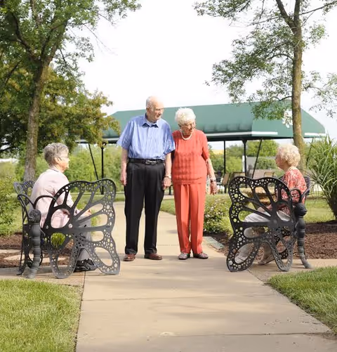 Two elderly women sitting on butterfly-shaped metal benches facing each other on a paved path in a garden area, while an elderly man and woman stand holding hands and looking at each other between the benches. Trees and a green canopy structure are visible in the background.
