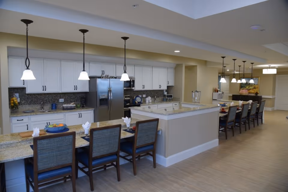 A spacious, modern kitchen and dining area with white cabinets, granite countertops, and stainless steel appliances. There are pendant lights hanging over the kitchen island and a long dining table with chairs set for a meal. The floor is light wood, and the walls are painted a soft beige color.