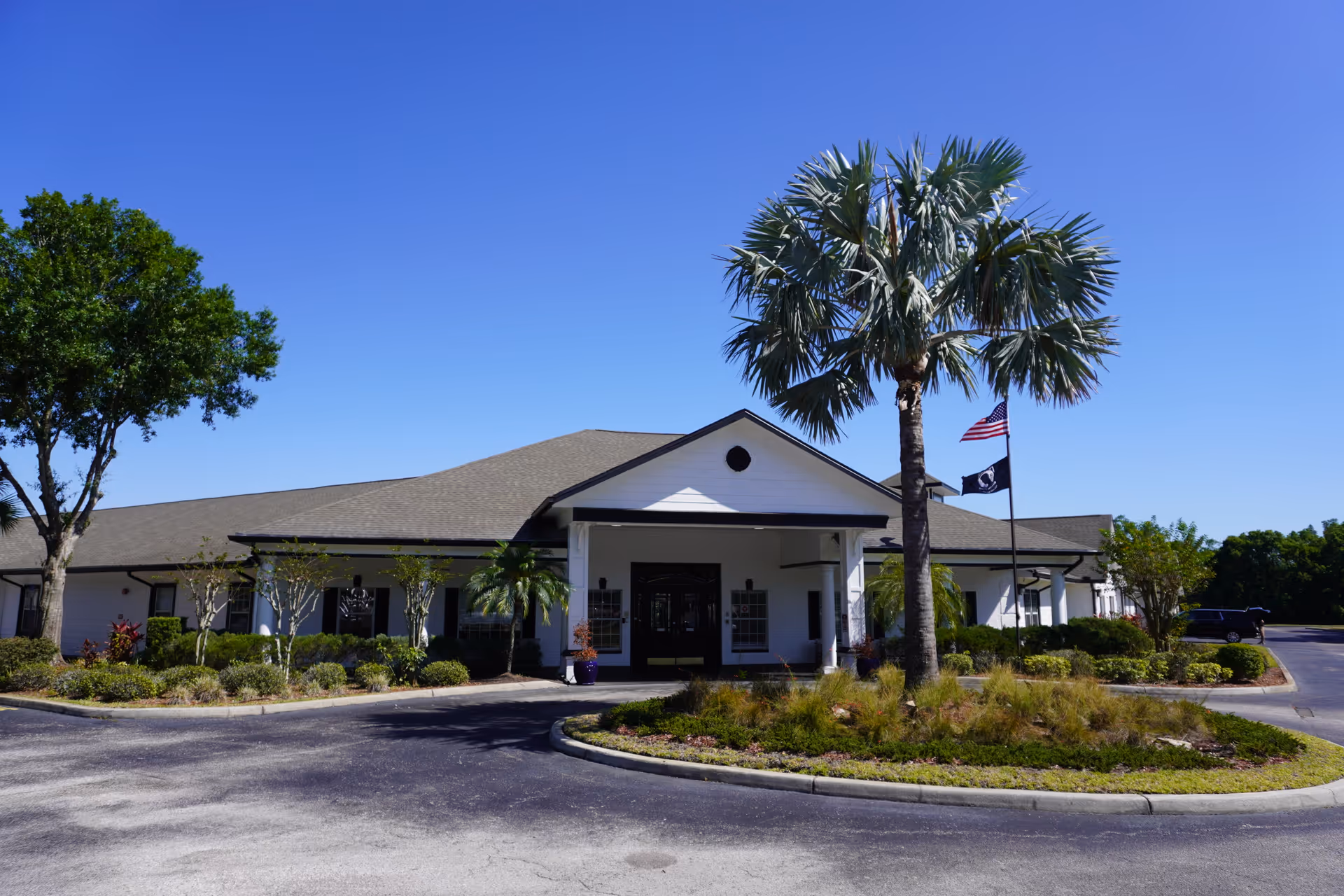 Exterior view of a single-story building with a gray roof and white walls, surrounded by landscaped greenery including palm trees and bushes. There is a circular driveway in front with an American flag and another flag on flagpoles near the entrance under a covered porch.