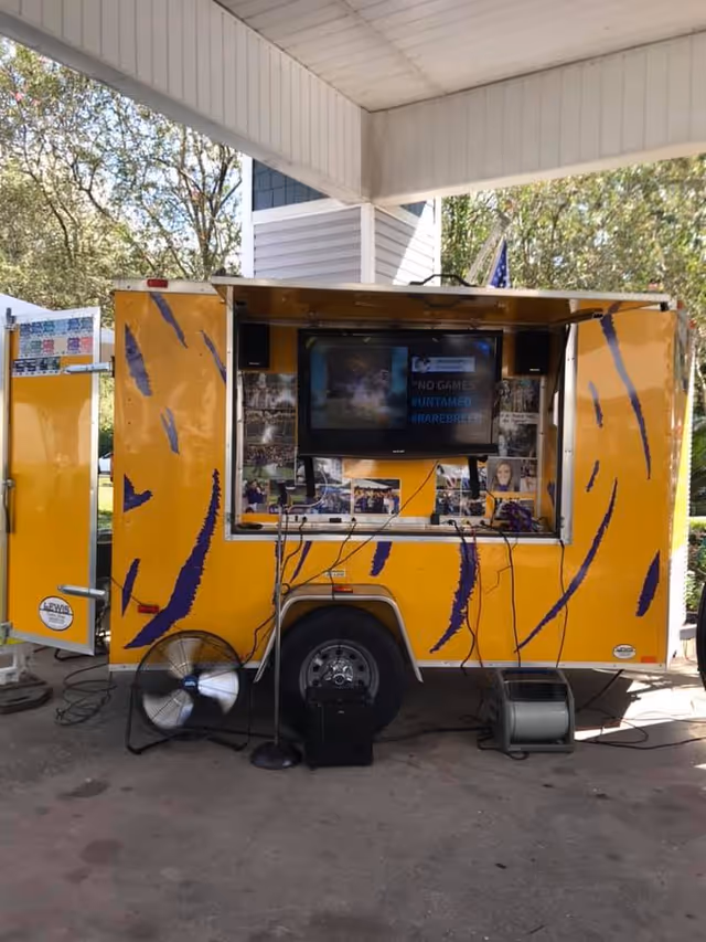 Yellow and purple mobile trailer booth with a mounted TV and audio equipment parked under a covered pavilion.