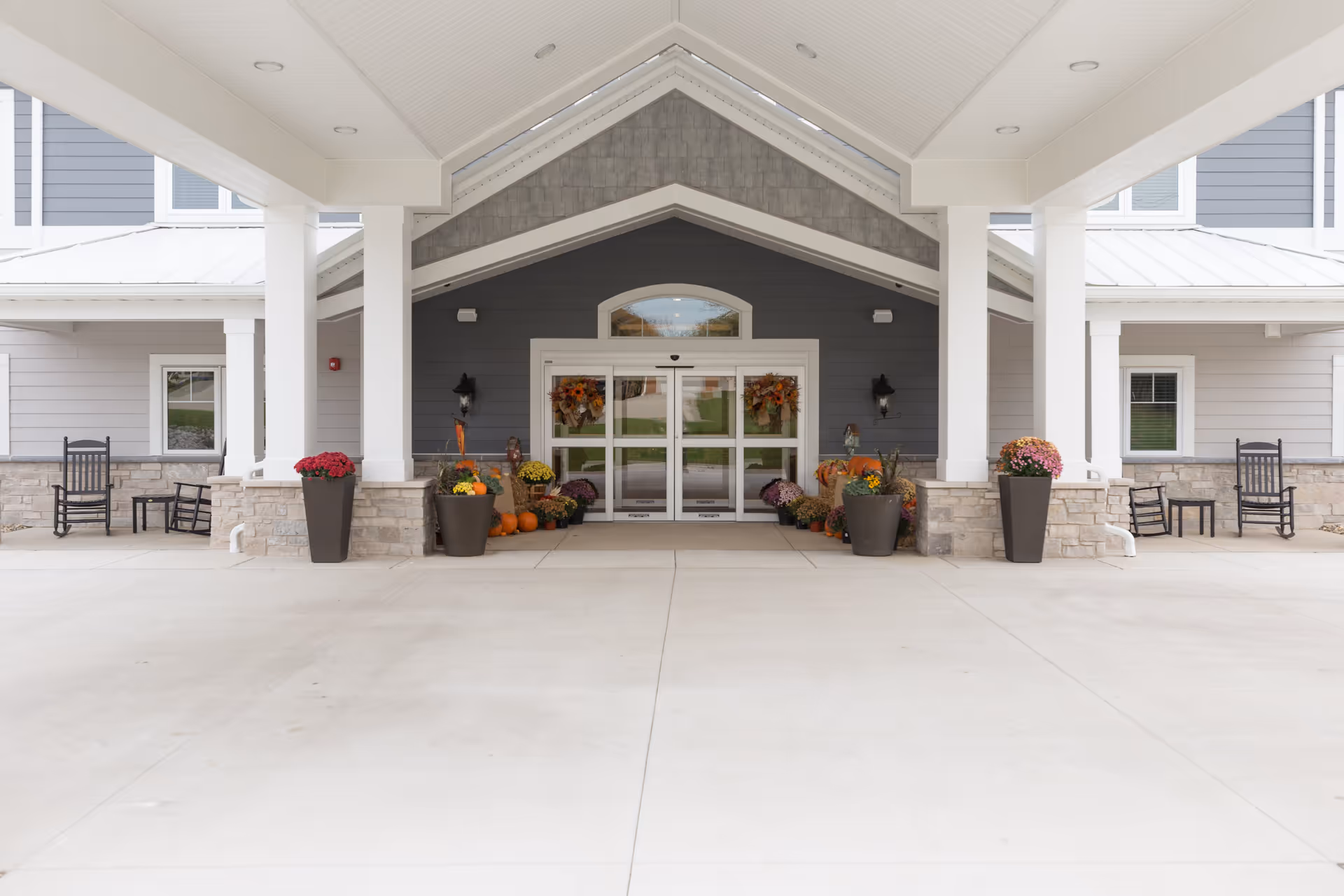 Covered entrance of Villas of Holly Brook Assisted Living & Memory Care in Brazil, IN, featuring automatic glass doors decorated with fall wreaths, large planters with flowers and pumpkins, and black rocking chairs on either side.