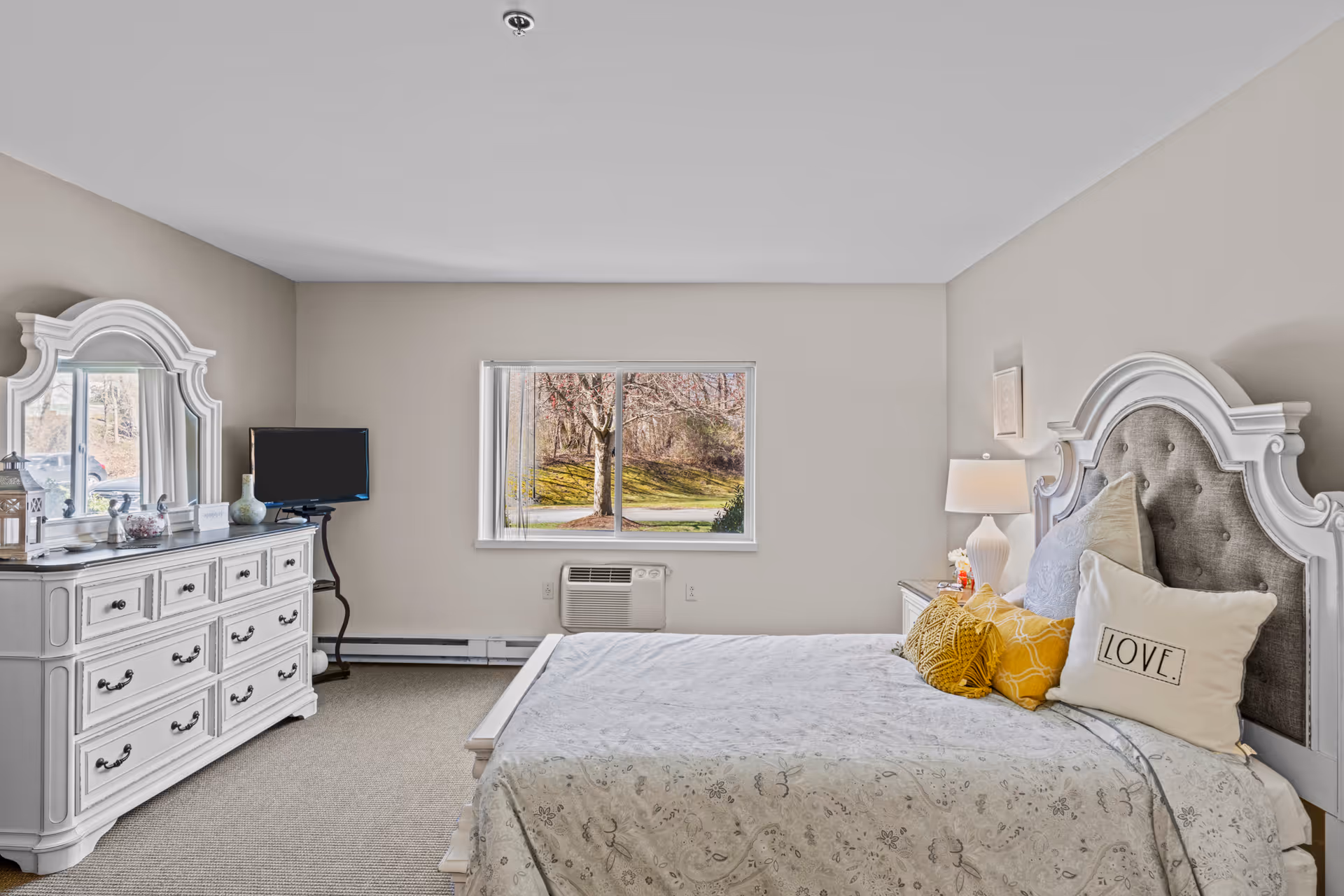 Bright furnished bedroom with an ornate white dresser and upholstered headboard, a made bed with decorative pillows, and a window showing trees outside.
