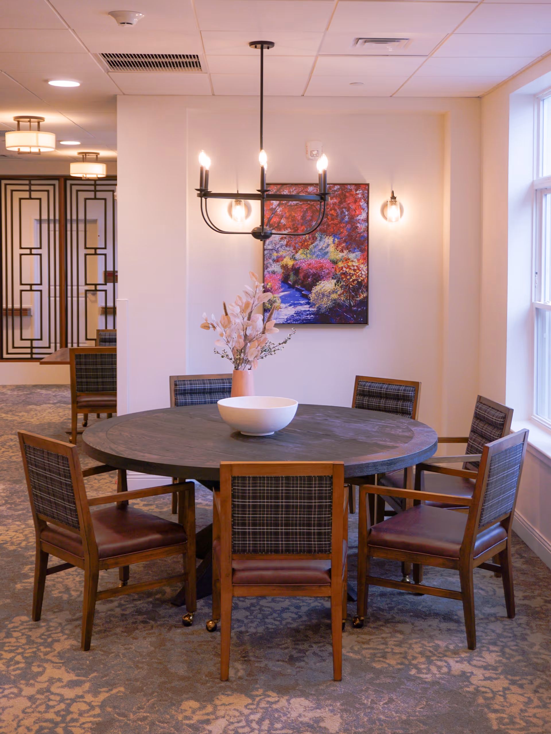 A round wooden dining table with six wooden chairs featuring plaid backs and leather seats. A white bowl and a vase with dried flowers are placed on the table. A modern chandelier with exposed bulbs hangs above the table. A colorful painting of a garden path is mounted on the wall behind the table. The room has carpeted flooring and a large window letting in natural light.