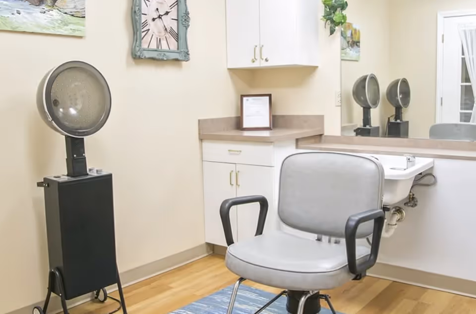 Interior view of a salon or barber area in a senior living facility, featuring a gray salon chair with black armrests, a hair dryer on a stand, a small sink, white cabinets with gold handles, a large mirror, and wall decorations including a clock and a painting.