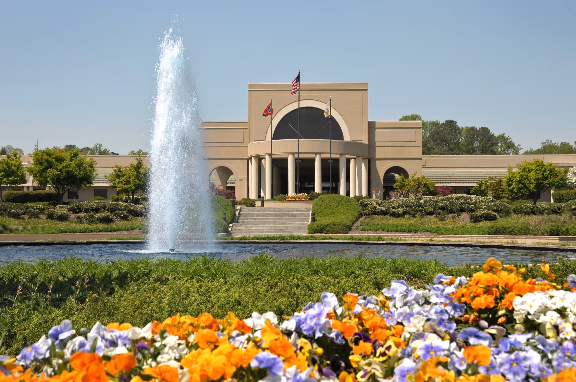 Front exterior view of a large beige building with a central entrance featuring columns and three flagpoles flying flags. In front of the building is a large water fountain surrounded by a pond, with colorful flowers and greenery in the foreground under a clear blue sky.