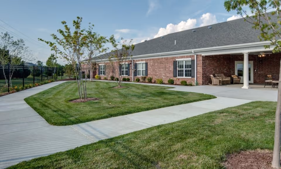 Brick single-story senior living building exterior with manicured lawn, walkways, and a covered patio with seating.