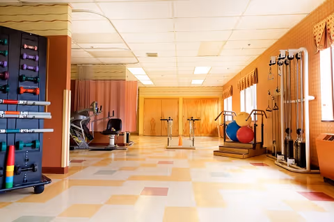 A rehabilitation exercise room with various fitness equipment including stationary bikes, resistance bands, exercise balls, and weight racks. The room has a tiled floor with a pattern of beige, yellow, and red squares, large windows with valances, and a drop ceiling with fluorescent lighting.
