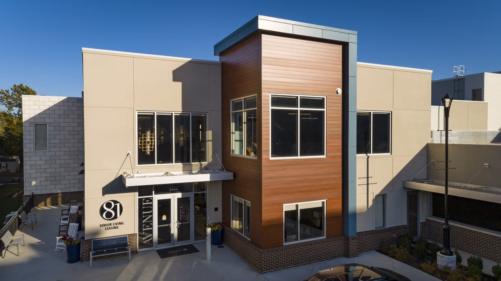 Exterior view of Avenue 81 Assisted & Independent Living facility showing the entrance with large windows, beige and wood-paneled walls, outdoor seating area with chairs, and a lamp post under a clear blue sky.