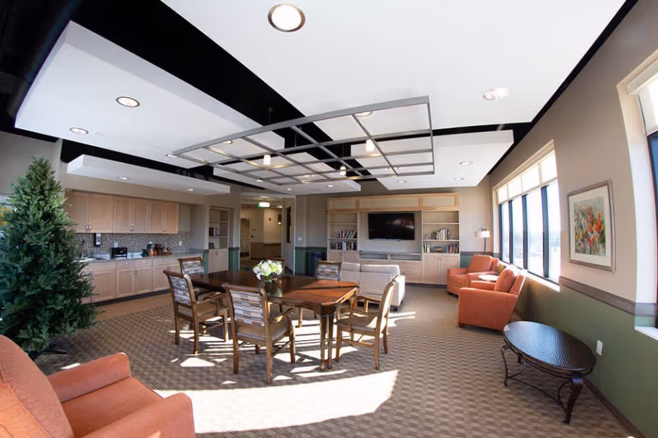 A bright and spacious common area in The Avalon of Bloomfield Township featuring a wooden table with six chairs in the center, surrounded by comfortable orange armchairs and a beige sofa. The room has large windows letting in natural light, built-in wooden shelves with a TV, and a kitchenette area with cabinets and a countertop. The ceiling has modern recessed lighting and a decorative grid structure.