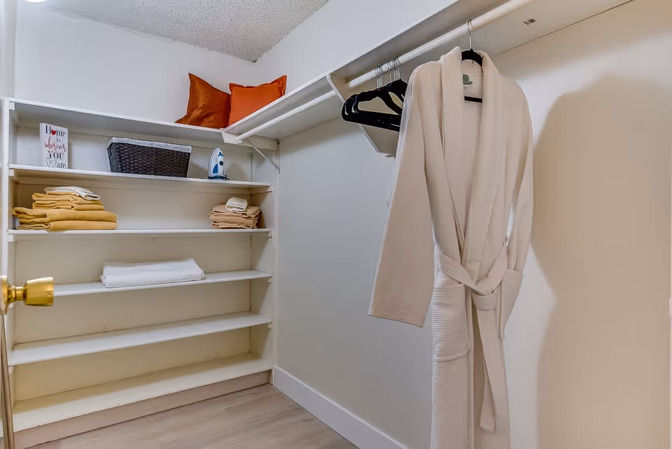 A small walk-in closet with white shelves holding folded towels, an iron, a basket, and two orange pillows. A white bathrobe hangs on a hanger from a rod on the right side.
