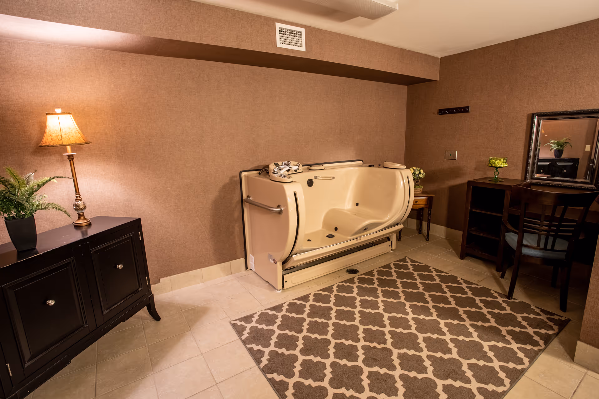 A room with beige walls and tiled floor featuring a beige walk-in bathtub with safety handles. To the left, there is a black cabinet with a lamp and a potted plant on top. To the right, there is a wooden vanity table with a mirror, a chair, and a small green decorative item. A patterned area rug is placed on the floor in front of the bathtub.