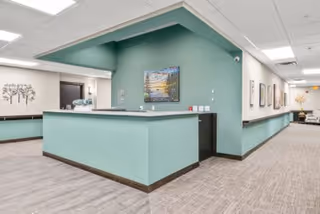 Reception area in a senior living facility with a light green front desk, green accent wall behind it, and a hallway extending to the right with framed artwork on the walls and seating visible in the distance.