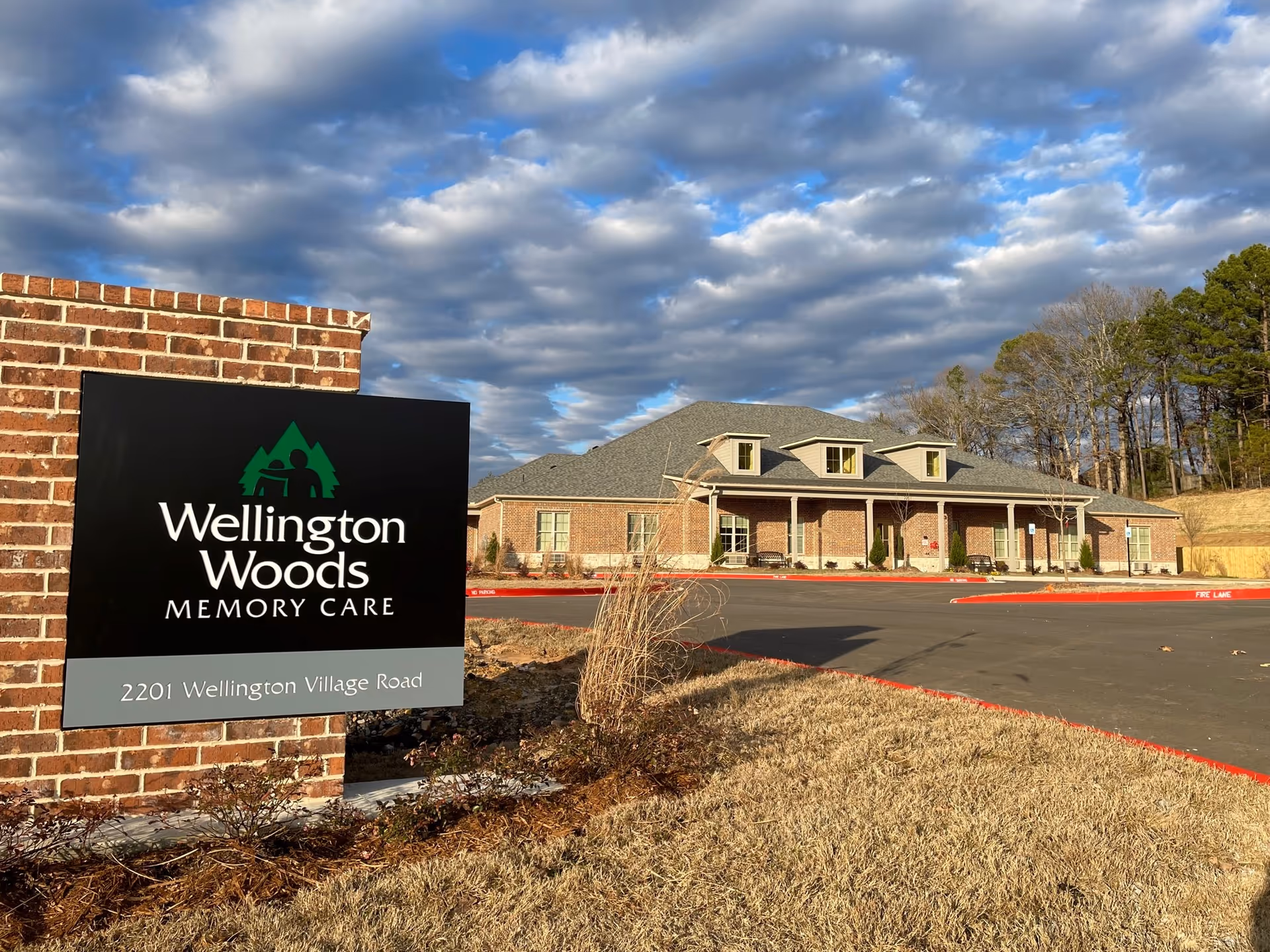 Exterior view of Wellington Woods Memory Care facility with brick sign, parking lot, and trees.