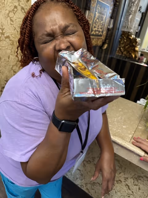 A woman with braided hair wearing a purple shirt and a black smartwatch is holding and biting into a partially opened silver foil snack package while standing indoors near a counter with decorative elements in the background.