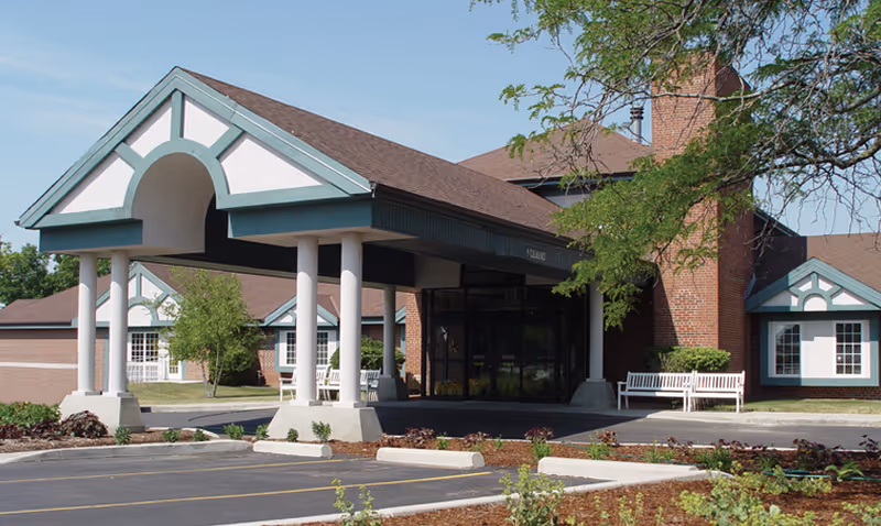 Front entrance of a brick senior living facility with a covered porte-cochère, parking area, and benches.