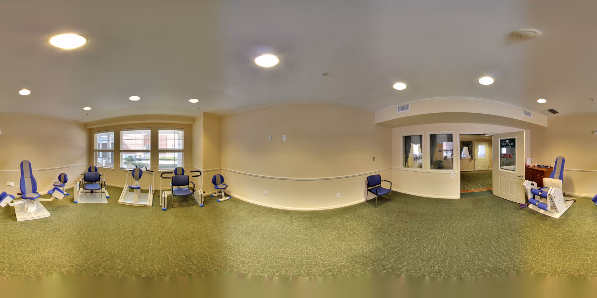 A spacious exercise room with several pieces of seated exercise equipment featuring blue and white padding. The room has beige walls, green carpet, and multiple ceiling lights. There are large windows letting in natural light and a door with a sign that reads 'Waltonwood Lakeside'.