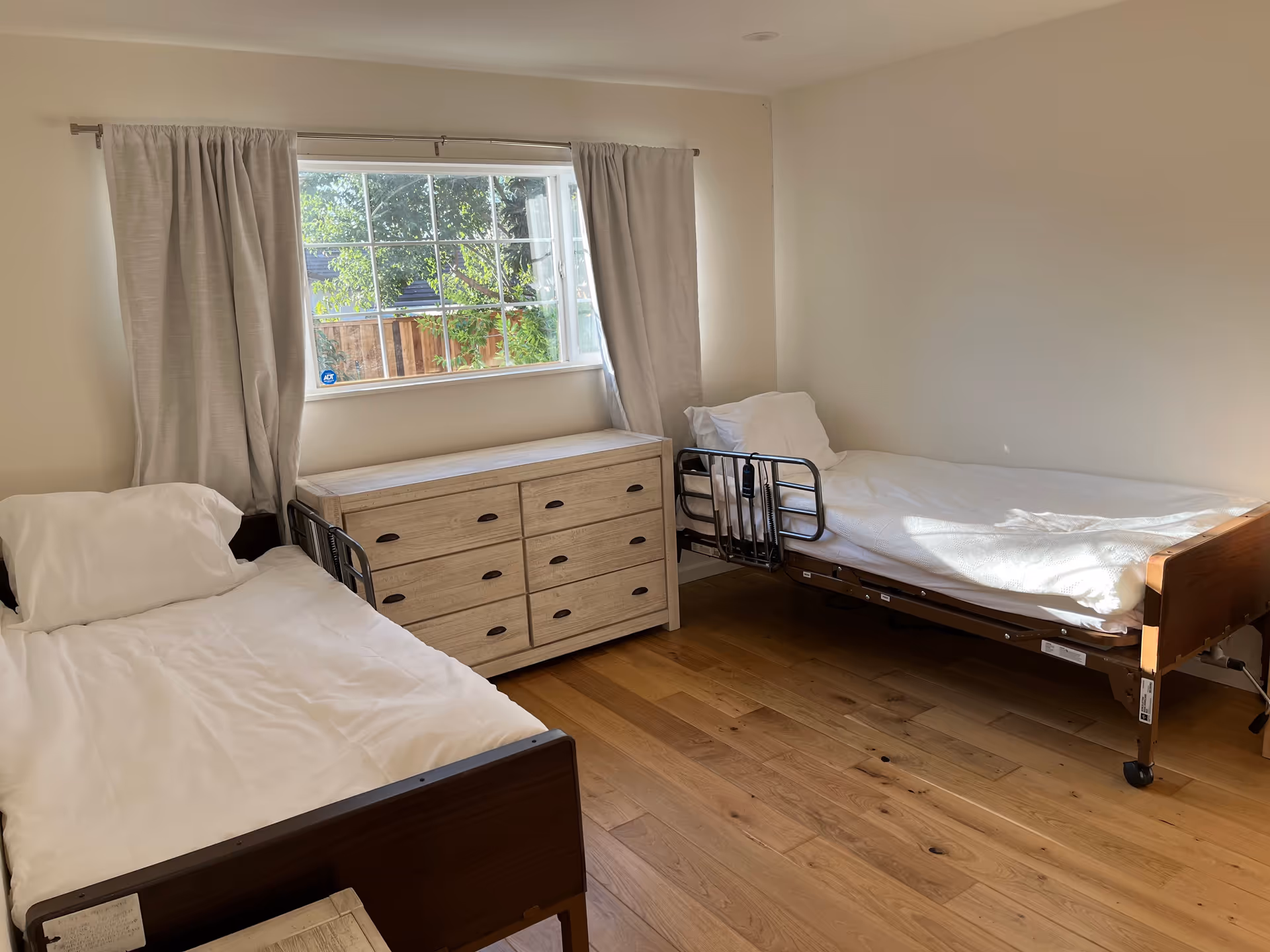 A simple, clean bedroom with two single beds, each with white bedding and pillows. Between the beds is a wooden dresser with six drawers. A large window with light-colored curtains lets in natural light, showing greenery outside. The floor is wooden, and the walls are painted white.