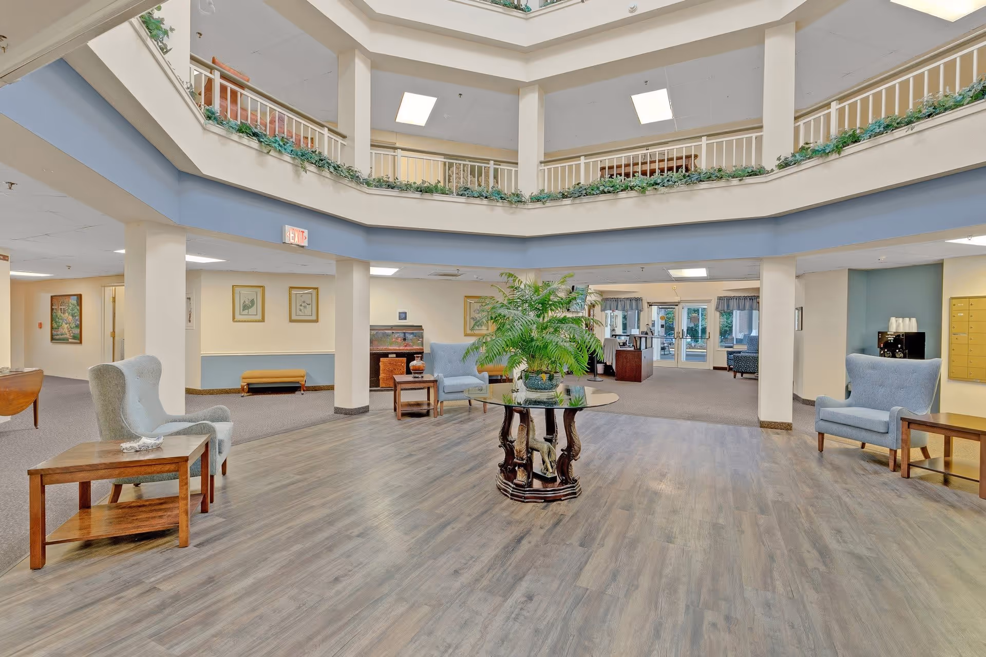 Spacious senior living community lobby with a circular open ceiling design featuring greenery along the upper railing. The area has light wood flooring, several comfortable blue armchairs with wooden side tables, framed artwork on the walls, and a central round table with a large potted plant. The lobby leads to other rooms and an entrance with glass doors.