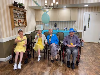 Four elderly individuals sitting in chairs and wheelchairs in a room with wooden flooring and beige striped wallpaper. They are holding yellow flowers and smiling. The room has a kitchen area in the background with cabinets, a chandelier, and some decorative items on shelves.
