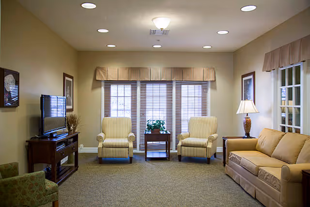 Bright communal living room with a sofa, two striped armchairs, a TV console, side table with lamp, and large windows with blinds.
