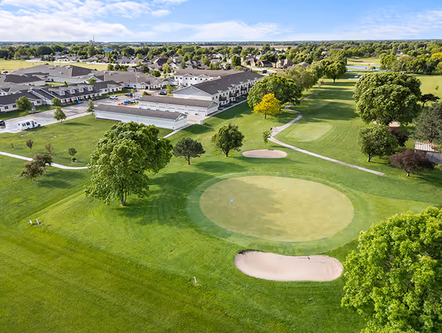 Aerial view of a senior living facility named Good Samaritan Society - Grand Island Village, showing multiple buildings surrounded by green lawns, trees, and a golf putting green with sand bunkers in the foreground under a partly cloudy sky.