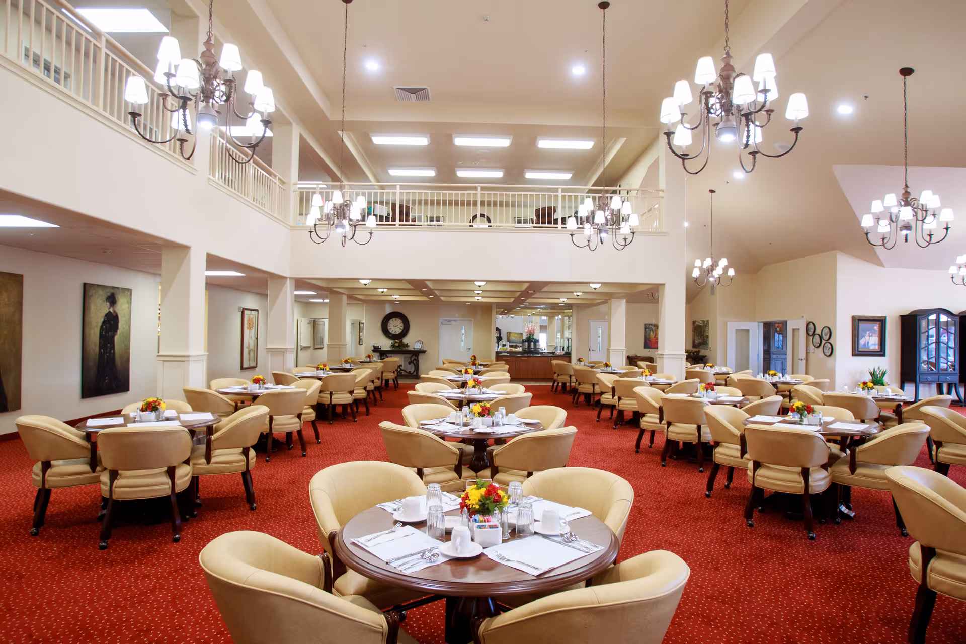 A spacious and elegant dining room with multiple round tables set with white tableware, glasses, and small floral centerpieces. The room features beige upholstered chairs, red carpet flooring, high ceilings with multiple chandeliers, and a balcony area overlooking the dining space. Artwork and decorative elements adorn the walls.