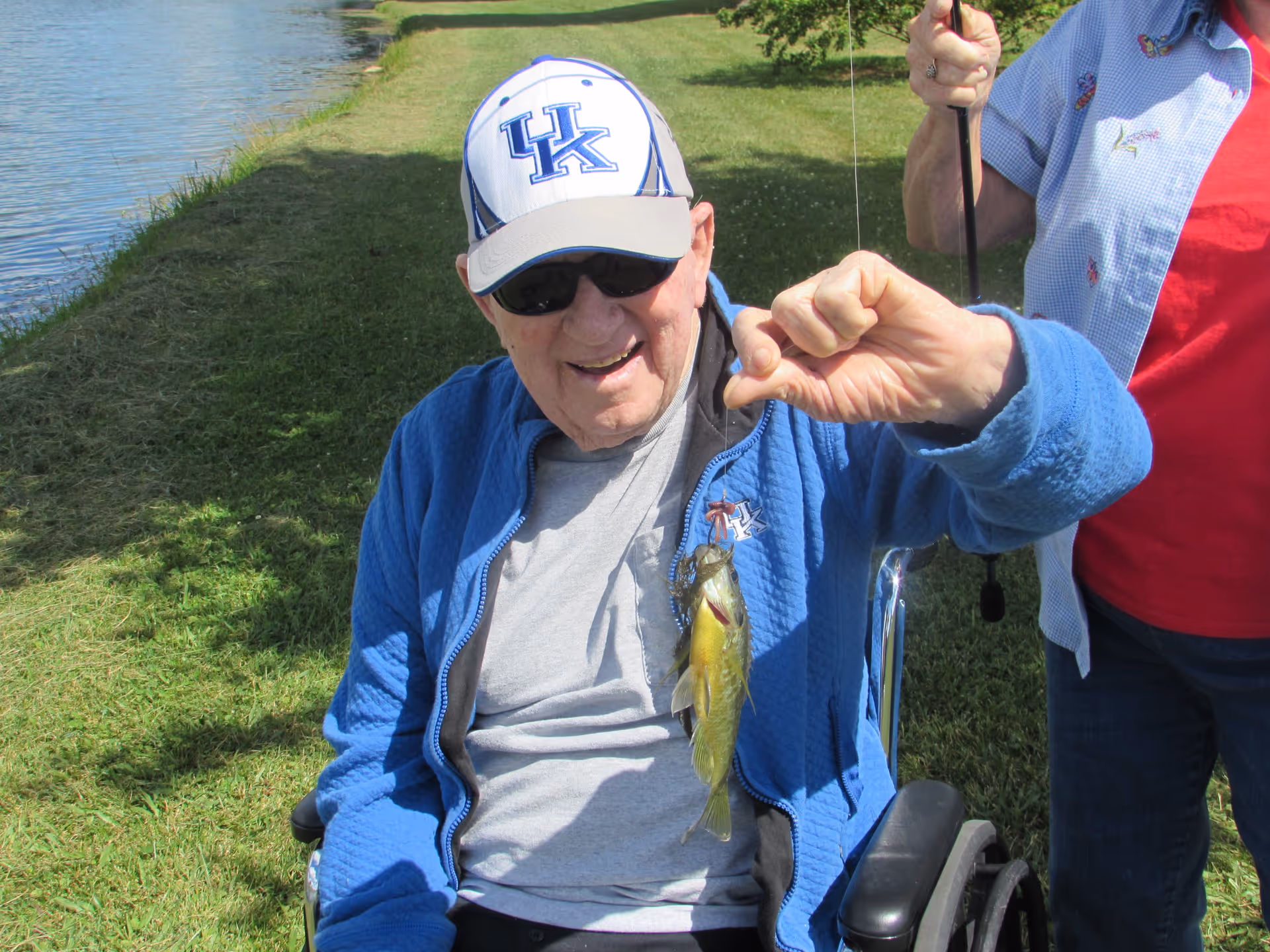An elderly man in a wheelchair wearing a blue jacket, gray shirt, sunglasses, and a white and blue UK baseball cap is holding up a small fish he caught while fishing by a grassy lakeside. A woman standing next to him is holding a fishing rod.