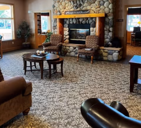 A cozy living room area with a stone fireplace as the focal point. Two patterned armchairs are placed in front of the fireplace, with a wooden coffee table and smaller side tables nearby. The room has large windows letting in natural light, a patterned carpet, and additional seating including a brown armchair and a black leather chair.
