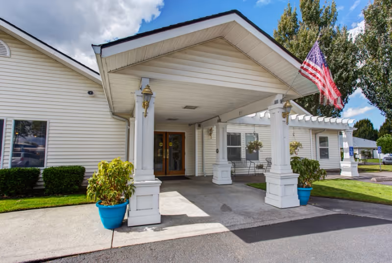 Entrance of a senior living facility named Evergreen Place with a covered driveway supported by white columns, two blue potted plants, an American flag, and a bench near the entrance under a clear blue sky with some clouds.