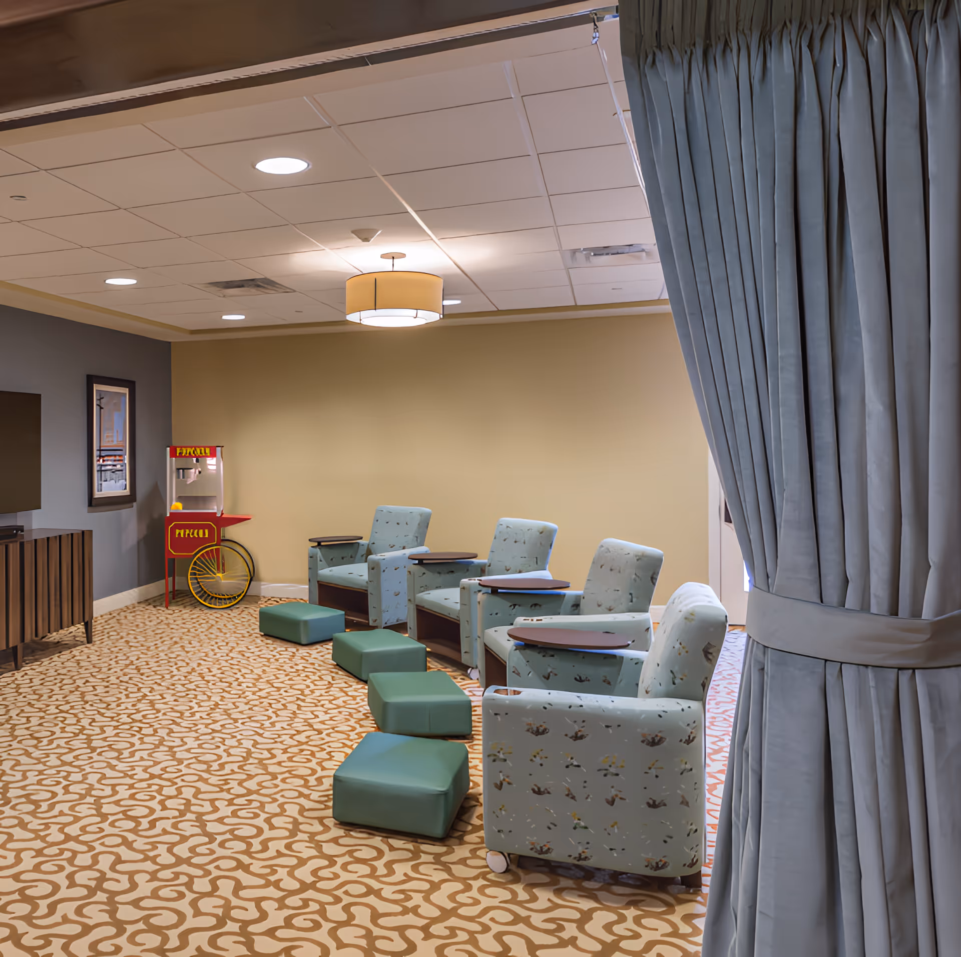 A cozy lounge area with four light blue armchairs each equipped with a small attached table, and four green footrests arranged in front of them. The floor is covered with a patterned carpet in beige and brown tones. A popcorn machine is placed against the far wall, and a large curtain is partially drawn to the right side of the image. The ceiling has recessed lighting and a central hanging light fixture.