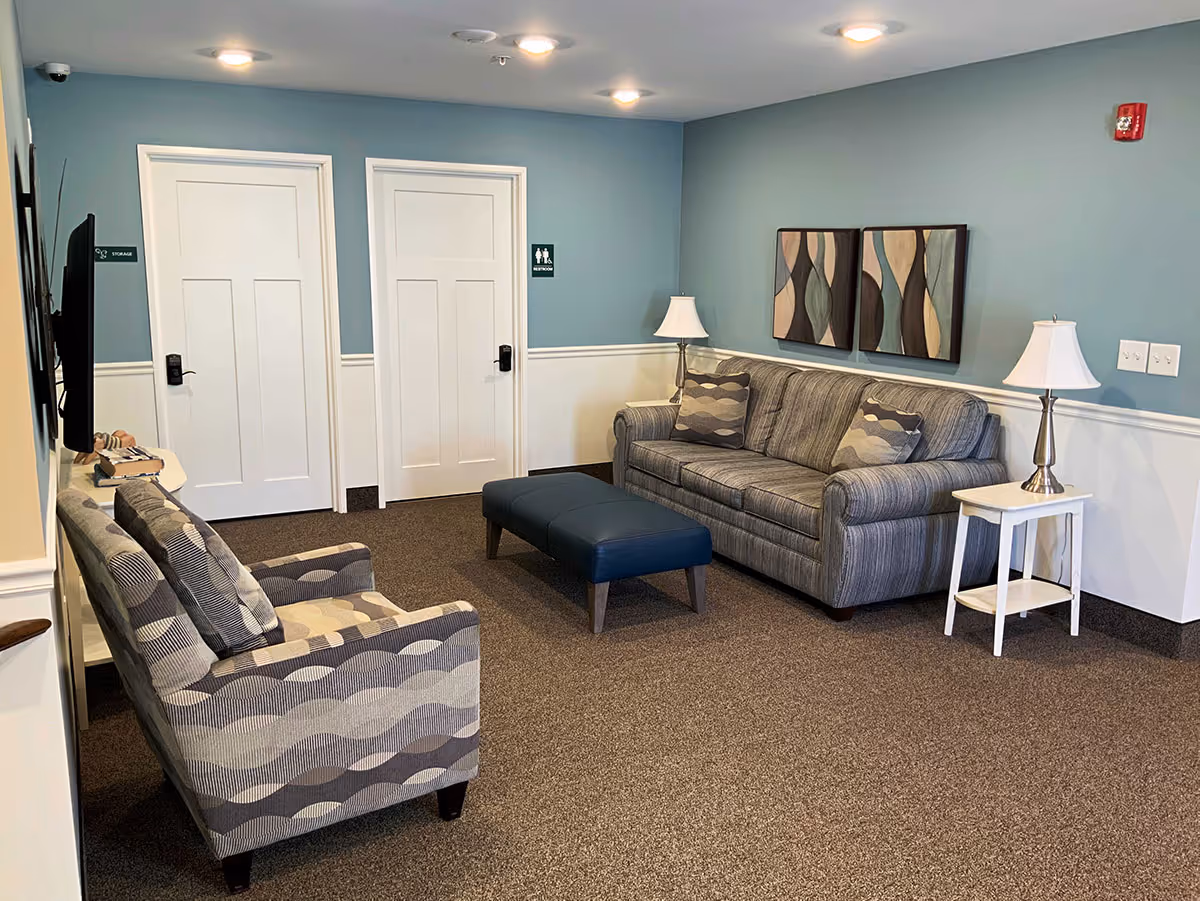 A cozy sitting area in a retirement living facility with a striped gray sofa, matching armchair, blue ottoman, two white side tables with lamps, abstract wall art, and two white doors labeled storage and restroom on a blue and white wall.